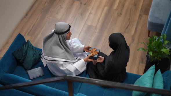 Top View Middle Eastern Husband Passing Gift to Surprised Wife Sitting on Couch at Home alt