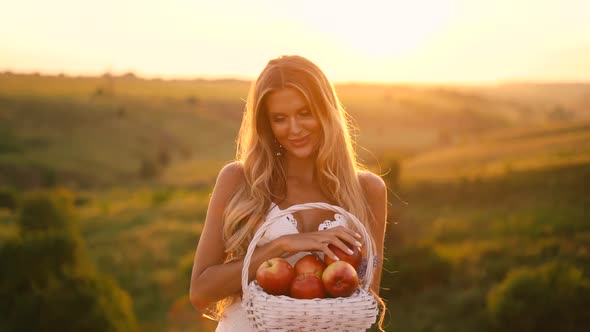 Beautiful sexy blonde girl in white dress posing in a field at sunset with a basket of fruit	 alt