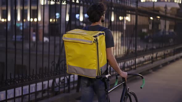 Delivery Man Walking with Bike and Yellow Bag By City Bridge Rear View Slow Motion alt