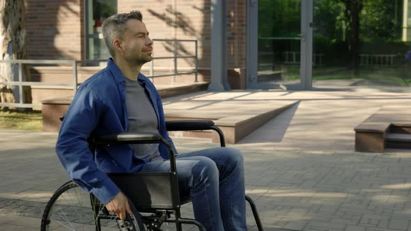 Side View of the Happy Disabled Man in Wheelchair is Riding in City Alone Moving Wheels with Hands alt