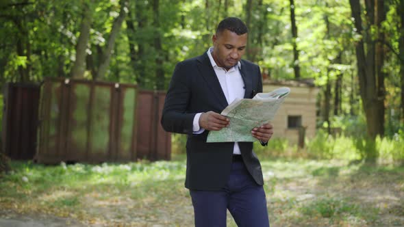 Confident African American Man Walking in Sunlight with Paper Map in Summer Park alt