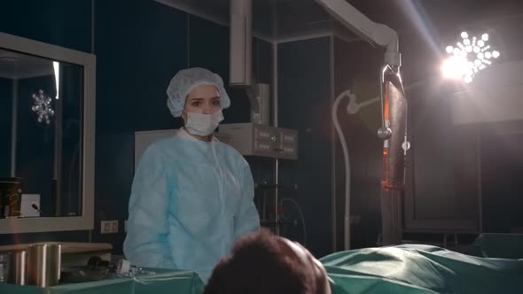 Female Surgeon in Sterile Gloves and a Robe Approaches the Patient on the Operating Table alt