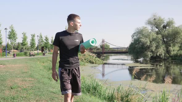 Young man walking along river in park with yoga mat in hands alt