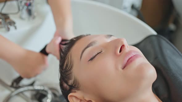 Caucasian young woman lying down and close eye on salon washing bed getting hair washed by stylist. alt