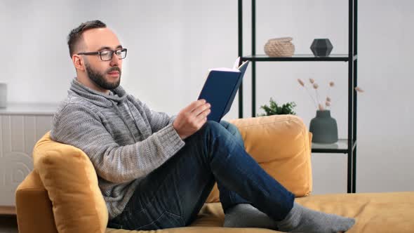 Handsome Guy Reading Paper Book Relaxing on Comfortable Yellow Couch alt