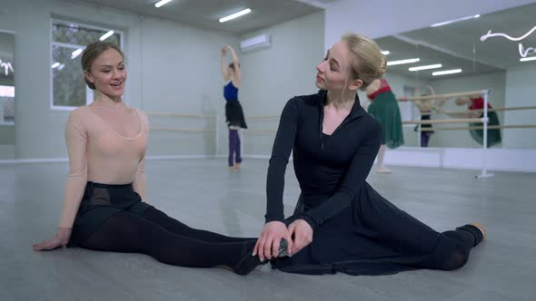 Smiling Ballerina Helping Friend Stretching Feet Sitting on Floor in Dance Studio alt
