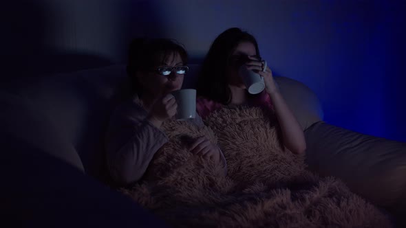 Mom and Daughter Sit in the Evening on the Couch with Mugs of Tea, Watch Tv and Laugh alt