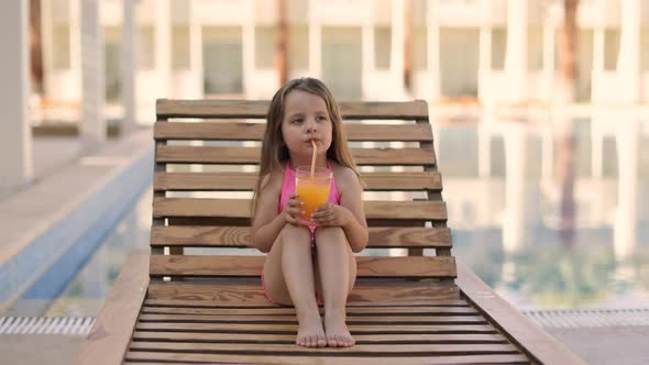 Portrait of Little Cute Girl in Pink Swimsuit with Orange Juice Glass Resting on a Wooden Lounge alt