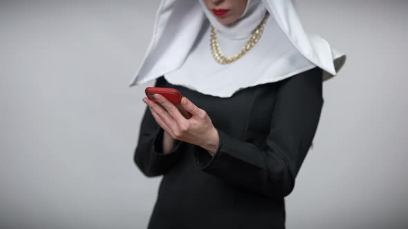 Unrecognizable Absorbed Woman in Nun Costume Texting on Smartphone Standing at Grey Background alt
