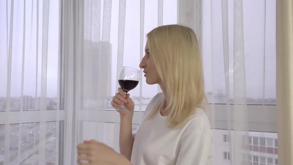 A beautiful girl holds a glass of wine and looks out the window. Wine tasting. alt