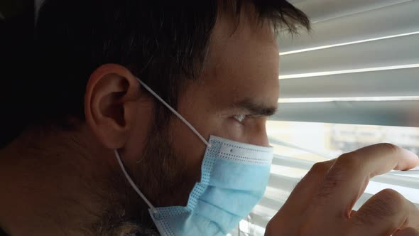 A Man Wearing a Medical Mask Approaches the Window Pulls Back the Blinds with His Hand and Looks Out alt
