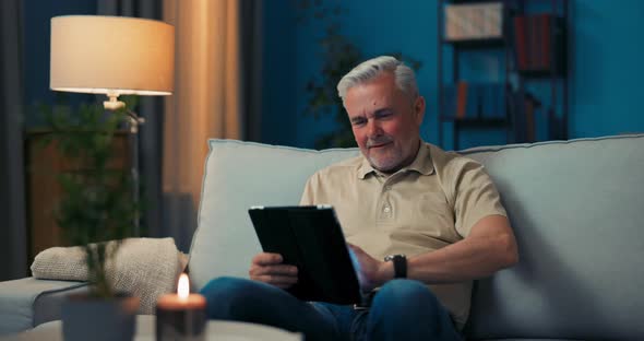Smiling Grayhaired Man Relaxing in Living Room on Sofa with Tablet alt
