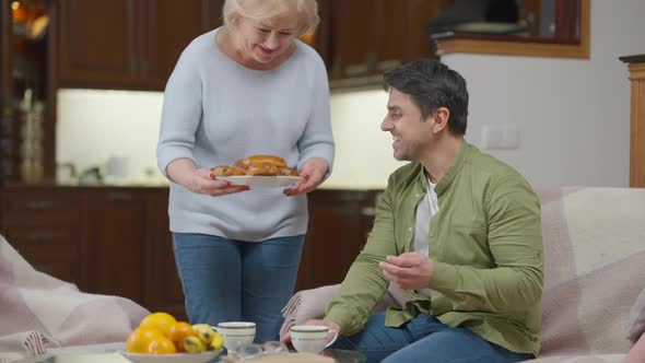Senior Mother Bringing Tasty Freshlybaked Buns for Son Sitting on Armchair in Living Room alt