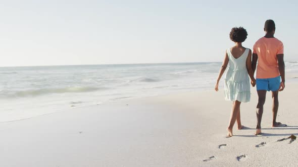 African american couple holding hands and walking on sunny beach alt