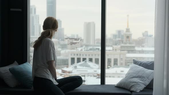 a woman sits on the windowsill and looks out the window at the city