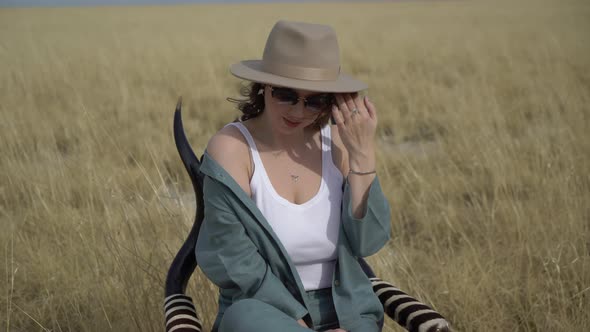 Young Woman in Hat Sits on Chair and Poses in the Savannah on a Dry Grass Field