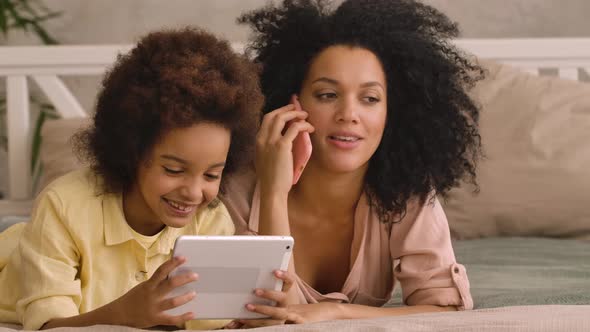 African American Woman is Talking on Smartphone Litle Girl is Playing on Tablet alt