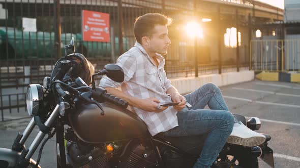 Young Man Motorcyclist with Smart Phone in Hands on Custom Motorcycle on Street alt
