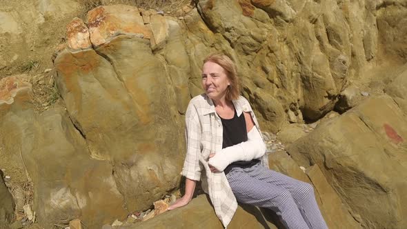 Senior Smiling Elderly Woman Relaxing Sitting on Rocky Coastline in Windy Day Suffering From Joint