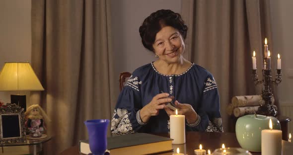 Portrait of Old Caucasian Woman in Blue Blouse Sitting at the Table with Candles and Shuffling Cards alt