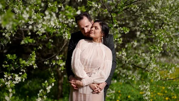 Portarit of Happy Lovers in Blooming Spring Garden Man is Embracing His Beautiful Wife alt