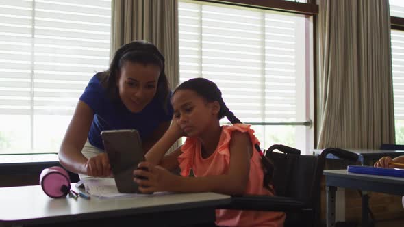 Diverse smiling female teacher helping schoolgirl sitting in wheelchair, using tablet alt