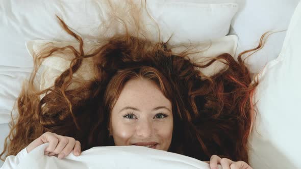 Attractive woman with red hair playfully looks out from under blanket