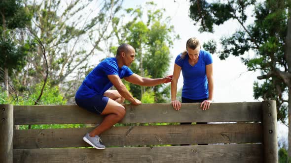 Male trainer assisting fit woman to climb over wooden wall during obstacle course 4k alt