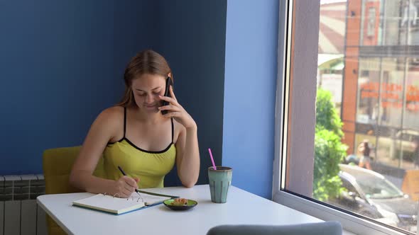 A Young Female Freelancer Sits in a Cafe Talking on a Mobile Phone and Writing Diary Entries alt