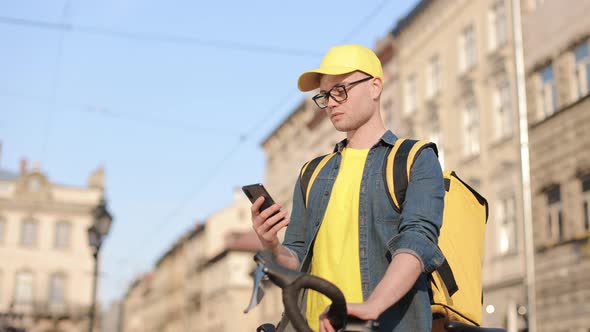 Portrait of a Happy Delivery Man Who is Sitting on a Bicycle and Texting on the Smartphone alt