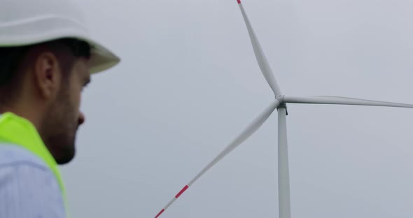 Bearded engineer in white helmet looks at rotating wind turbine.