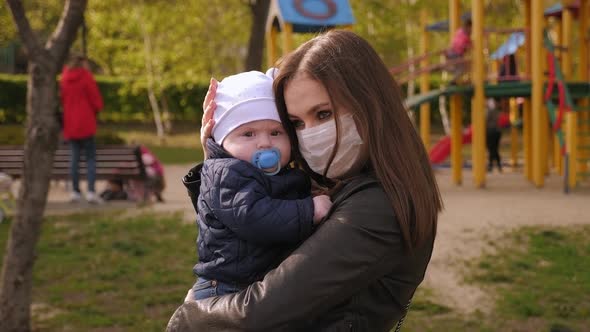 A Mother in a Medical Mask Hugs Her Little Son in a Park on a Crowded Playground alt