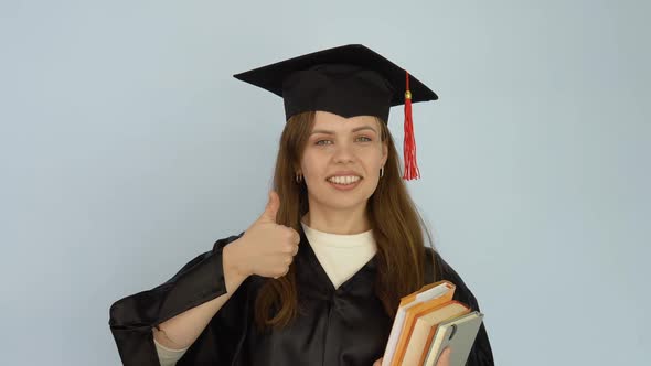 A Young Caucasian Woman in a Black Robe and a Master's Hat Stands Straight Holding Textbooks and alt