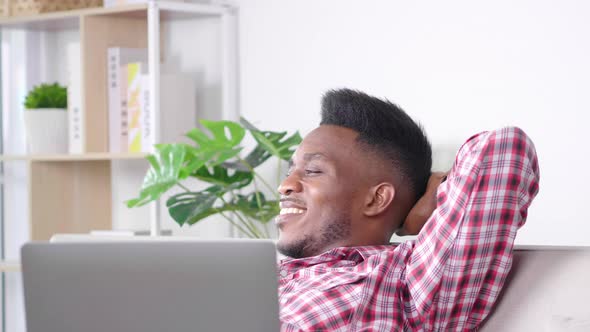 Happy African man feels relaxing on sofa in break time from work, putting laptop alt