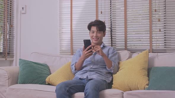 In his living room, an Asian man is on the phone, listening and conversing on his smartphone. alt