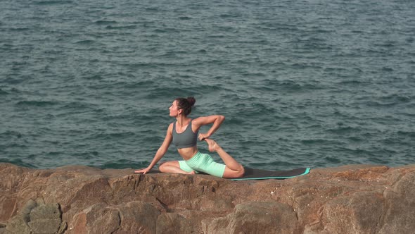 Young Woman Does Yoga in Pigeon Pose on Big Rock Against Sea