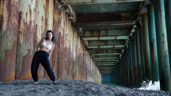 a Barefoot Woman Dances Under the Pillars of the Bridge Against the Background of the Incoming Waves alt