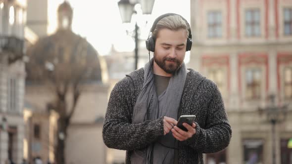 A Happy Bearded Man is Listening to Music with Headphones and Scrolling Through Social Networks on