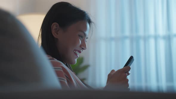 Asian beautiful woman sit on sofa and chat on mobile phone at night. alt