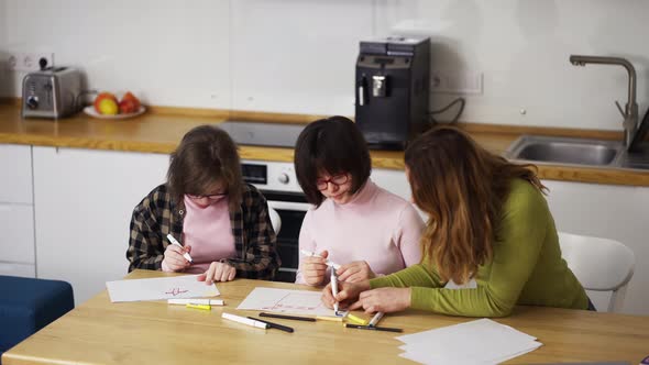 Two Girls with Down Syndrome Draw at the Kitchen Next to the Teacher or Mother Together alt
