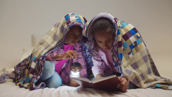 Elementary Age Sisters Reading Book Under Blanket alt