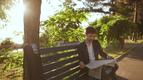 Slow Motion of Handsome Young Business Man in Suit Sitting on Bench Resting in Green Park at Sunset