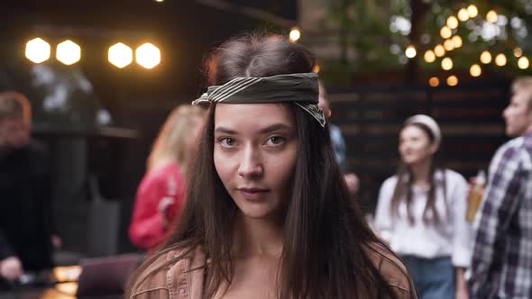Young Woman in a Bandanna with Long Hair Posing to the Camera while Her Friends Dancing alt