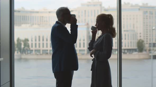 Side View of Senior Couple Drinking Coffee Standing Near Panoramic Window and Talking alt