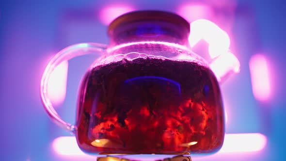 Transparent Teapot with Tea Leaves Boils on Gas Burner Against Neon Backlight alt