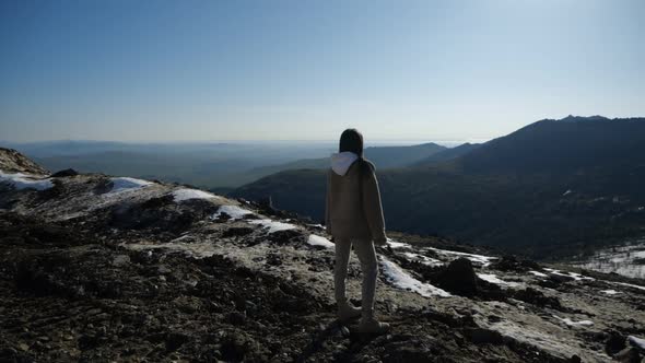 A Young Girl in a Beige Hooded Jacket Stands in the Mountains
