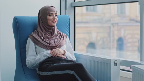 Smiling Dreamy Arabic Girl in Hijab Looking Out Window Sitting in Armchair Waiting for Exam or alt