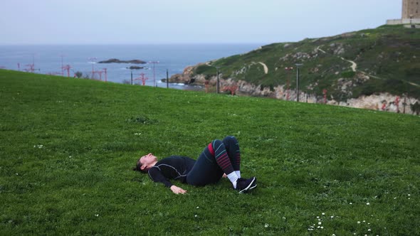a Girl in Blue Leggings Lying on Green Grass and Doing Exercises with Rubber for the Buttocks alt