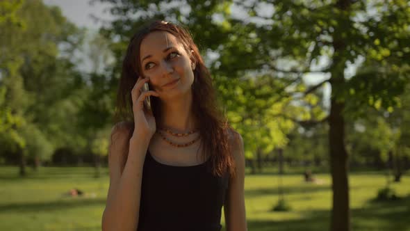 Smiling Young Lady Standing at the Park Outdoors Talking By Phone alt