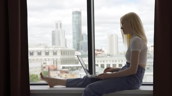 Young Joyful Woman Talking on a Video Call Using a Laptop While Sitting on a Windowsill By a Large